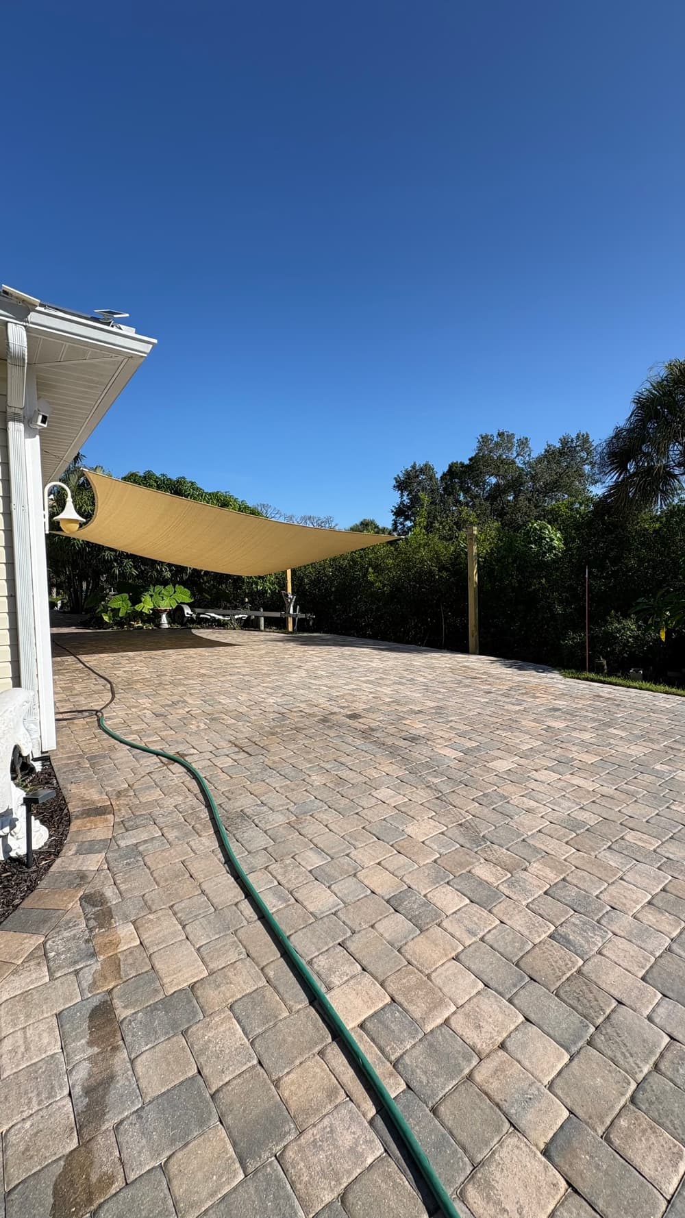 Paver patio with shade sail under blue sky and lush greenery in background.